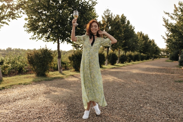 Trendy lady with red hairstyle in hat long yellow dress and white sneakers smiling and holding glass with wine on background of road