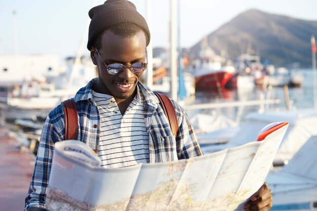 Trendy looking African American tourist with backpack in hat and sunglasses studying directions using city guide while exploring sights and landmarks of resort town