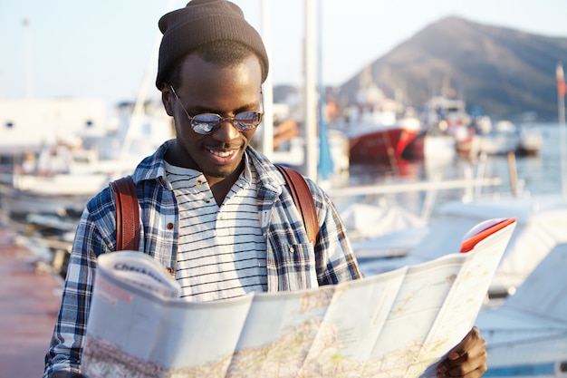 Модно выглядящий афроамериканский турист с рюкзаком в шляпе и солнцезащитных очках изучает направления, используя путеводитель по городу, исследуя достопримечательности и достопримечательности курортного города