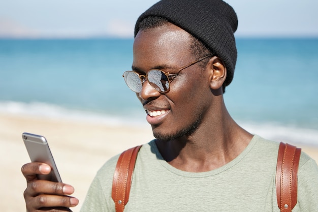 Giovani hipster dalla pelle scura alla moda con tonalità delle lenti specchiate e copricapo che leggono il messaggio o sfogliano i newsfeed tramite i social network, apprezzano i post e lasciano commenti mentre si riposa sulla spiaggia