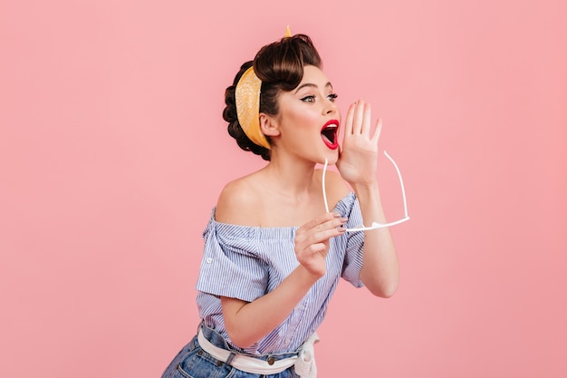 Trendy pinup girl screaming on pink background. Lovable young woman in retro outfit expressing excitement.