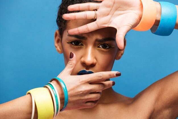 Trendy portrait of scared or thrilled mulatto woman with trendy makeup and accessories covering face with hands, over blue wall