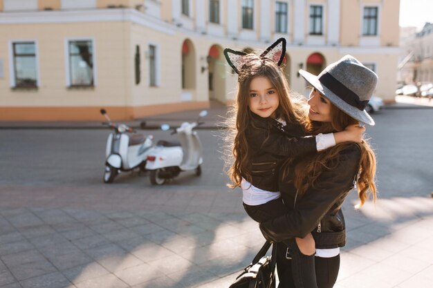 Trendy shapely woman with bag carrying curly brunette girl, smiling at camera. Portrait of young mother in stylish hat spending time with lovely daughter in weekend, going to shopping