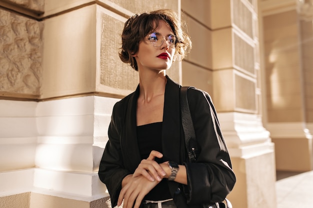 Trendy woman with wavy hairstyle and red lips posing outside. Brunette woman in dark jacket and glasses looks away outdoors.