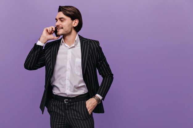 Модный молодой мальчик с брюнеткой в светлой рубашке и классическом полосатом костюме кладет одну руку в карман, разговаривает по мобильному телефону и смотрит в сторону Бизнес-концепция изолирована на фиолетовом фоне