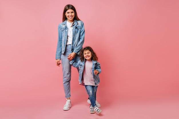 Trendy young woman holding hands with curly daughter. Studio shot of attarctive mom and preteen kid dancing together.