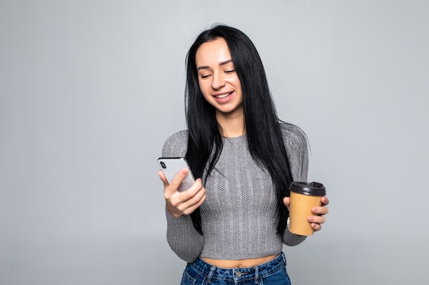 Trendy young woman standing chatting on a mobile phone while holding a mug of takeaway coffee in her other hand, isolated on gray wall