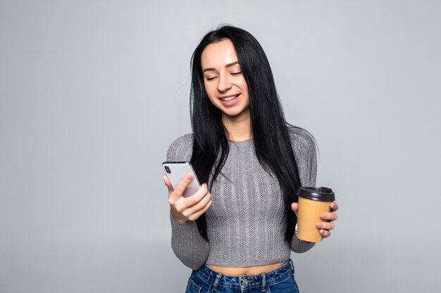 Trendy young woman standing chatting on a mobile phone while holding a mug of takeaway coffee in her other hand, isolated on gray wall