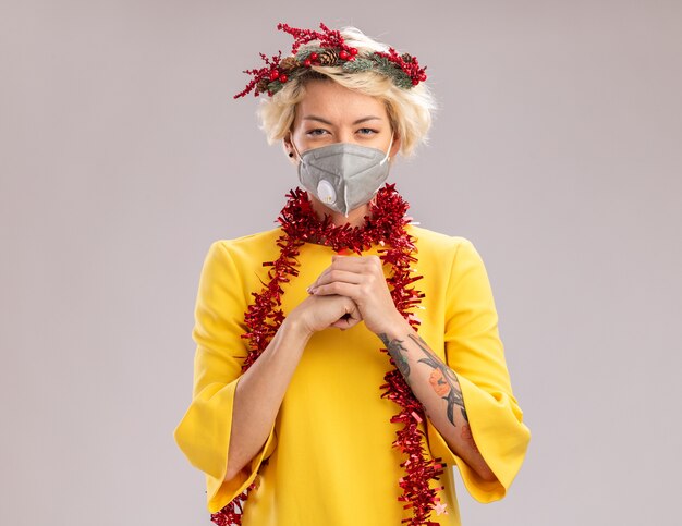 Tricky young blonde woman wearing christmas head wreath and tinsel garland around neck with protective mask looking at camera keeping hands together isolated on white background with copy space