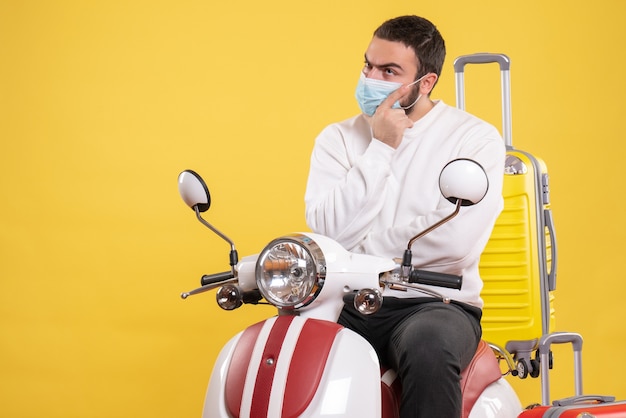 黄色のスーツケースを乗せたオートバイに座っている医療マスクを着た混乱した男との旅行のコンセプト