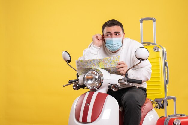 Trip concept with curious young guy in medical mask sitting on motorcycle with yellow suitcase on it and holding map