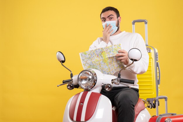 Концепция поездки с удивленным парнем в медицинской маске, сидящим на мотоцикле с желтым чемоданом и держащим карту на желтом