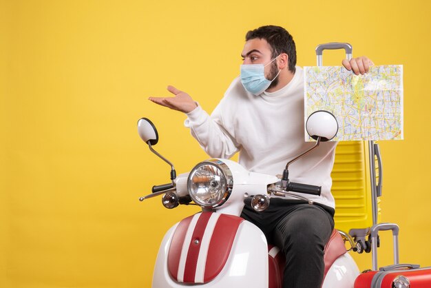 Trip concept with surprised guy in medical mask sitting on motorcycle with yellow suitcase on it and showing map on yellow