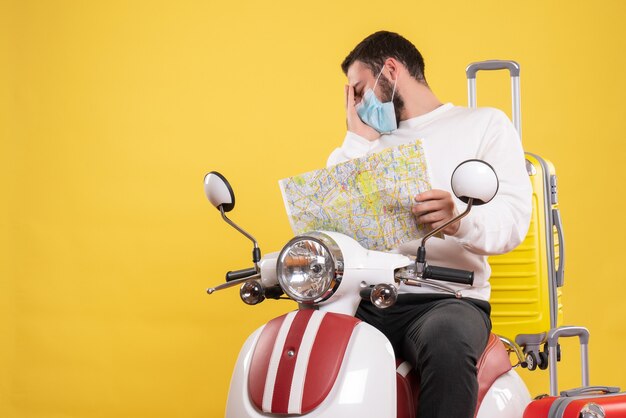 黄色いスーツケースを乗せたバイクに座り、頭痛に苦しんでいる地図を持った医療マスクを着た問題のある男との旅行のコンセプト