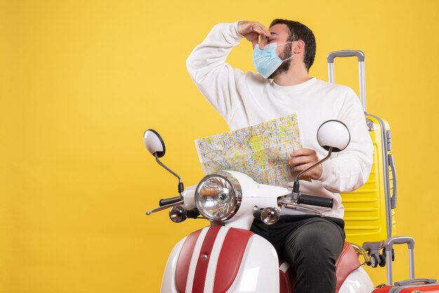 Trip concept with young guy in medical mask sitting on motorcycle with yellow suitcase on it and holding map making bad smell gesture