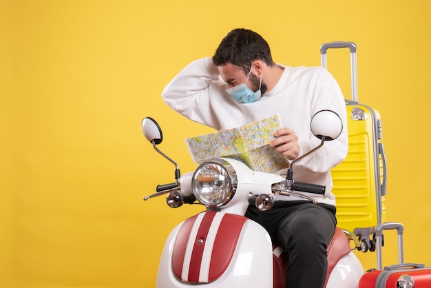 黄色いスーツケースを乗せたバイクに座り、頭痛に苦しんでいる地図を持った医療マスクを着た若い男との旅行のコンセプト