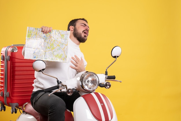 Concetto di viaggio con un giovane ragazzo seduto in moto con la valigia che soffre di infarto su di essa su giallo