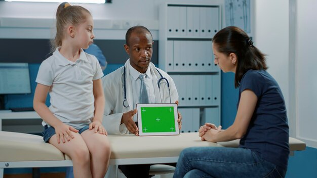 キャビネット内のタブレットに水平方向の緑色の画面を表示している小児科医の三脚ショット。空白のコピースペースと孤立したモックアップの背景が表示されているクロマキーテンプレートを見ている男性と患者。