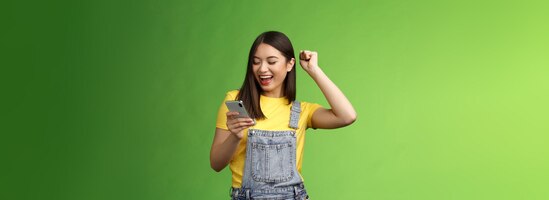 Trionfante ragazza asiatica gioiosa carina tenere pompa pugno smartphone celebrare buone notizie leggere buone notizie mes