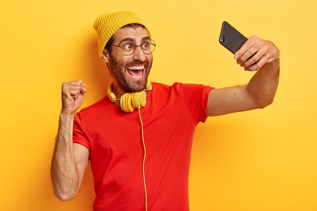 Triumphing glad man celebrates success, makes photo of himself, takes selfie, shoots video, wears hat, t shirt and spectacles isolated over yellow background. People