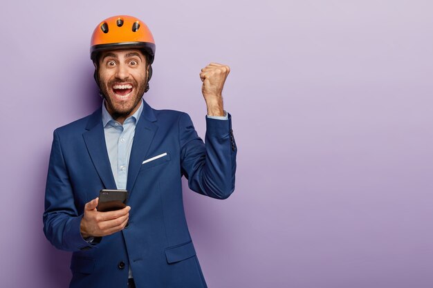 Triumphing happy engineer holds mobile phone, raises clenched fist, uses phone, rejoices being on construction site, wears formal suit and orange helmet. Young architect gets message on cellular