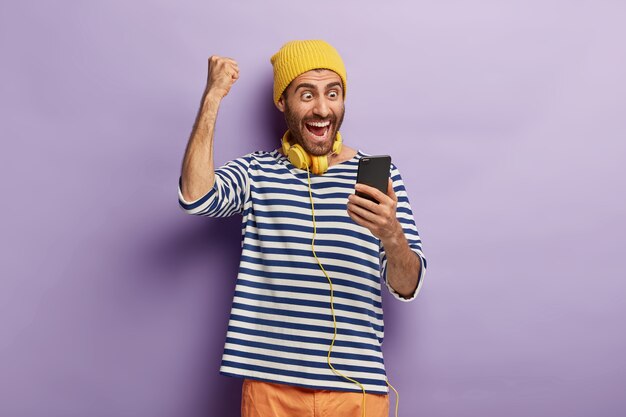 Triumphing happy guy raises clenched fist, celebrates winning lottery, gets message of confirming holds mobile phone, browses social media, wears yellow hat, striped jumper, always stays in touch