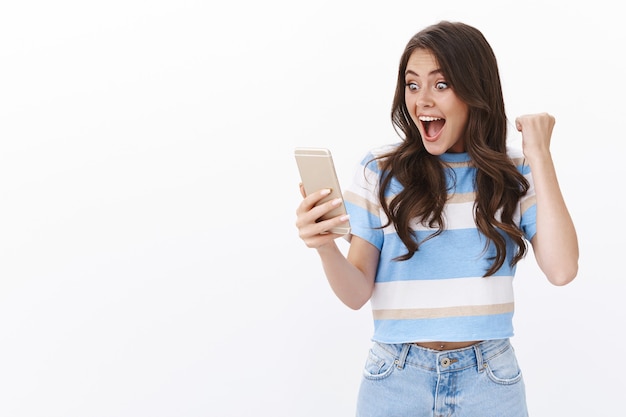 의기양양한 놀란 여성은 긍정적인 소식을 받고 스마트폰으로 문자 메시지를 읽는 동안 주먹을 휘두르며 눈을 뜨고 성공을 달성하고 온라인 복권에 당첨되었습니다.