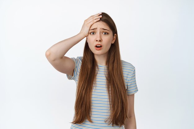 Troubled, anxious young woman holding hand on forehead with alarmed, upset face, forgot or remember smth bad, standing distressed on white