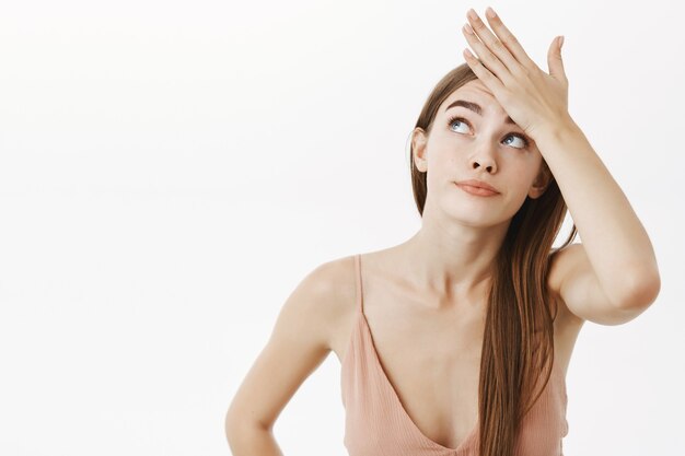 troubled beautiful caucasian female in beige evening dress holding hand on forehead and peeking up with intense bothered smirk being displeased with tiresome duties over gray wall