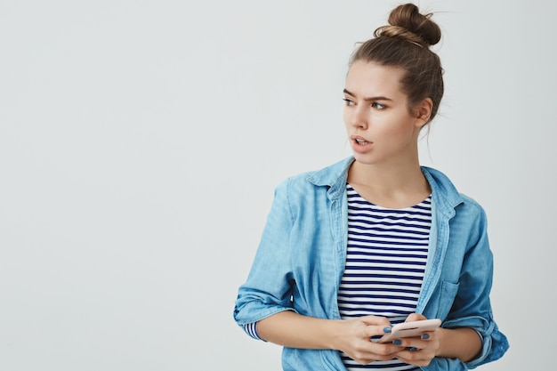 Free photo troubled concerned young caucasian female wearing hairbun, loose shirt, looking aside perplexed worried frowning receiving disturbing unpleasant message reacting troublesome news, holding smartphone