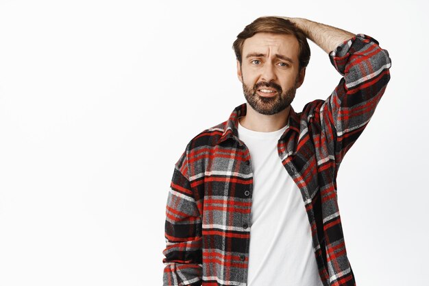 Troubled and confused bearded man holding hand on head staring puzzled and sad at camera white background