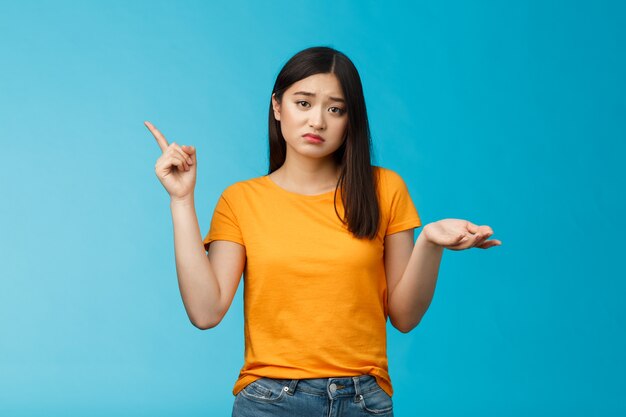 Troubled cute upset asian girl have problem asking your help, pointing upper left corner shrugging unsure, look frustrated and sad, questioned answer, cannot deal problem, stand blue background