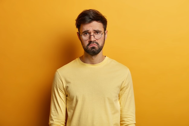 Troubled displeased bearded man frowns face, feels sad, distressed and upset, being bored sitting on quarantine, unhappy to miss good chance, dressed casually, isolated over yellow wall.