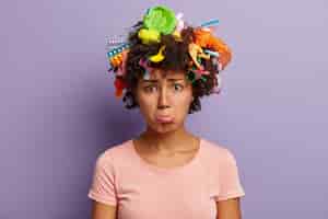 Бесплатное фото Обеспокоенная недовольная женщина поджимает нижнюю губу, собирает пластиковый мусор, носит повседневную футболку, экологически чистая, расстроена серьезной экологической проблемой, имеет мусор в кудрявых волосах, изолированных на фиолетовой стене