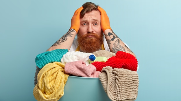 Troubled ginger young man keeps hands on head, looks desperately, wears protective gloves, has tattooed arms, sorrowful facial expression, stands against blue wall. People, domestic work.