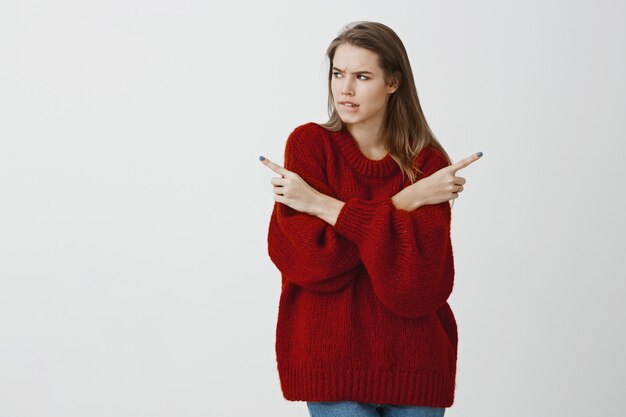 問題のある女の子がタフなディシソンを作ります。緩い赤いセーター、手を交差し、さまざまな方向を指して、神経質に唇をかむ、灰色の壁に何かを求めている疑わしい魅力的な女性