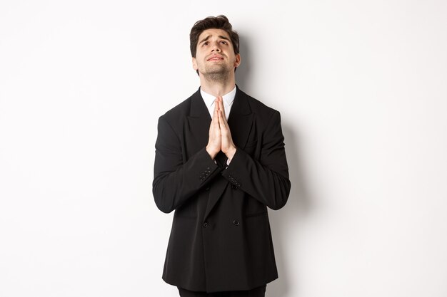 Troubled and hopeful man in black suit begging god, pleading and looking up, need help, standing over white background