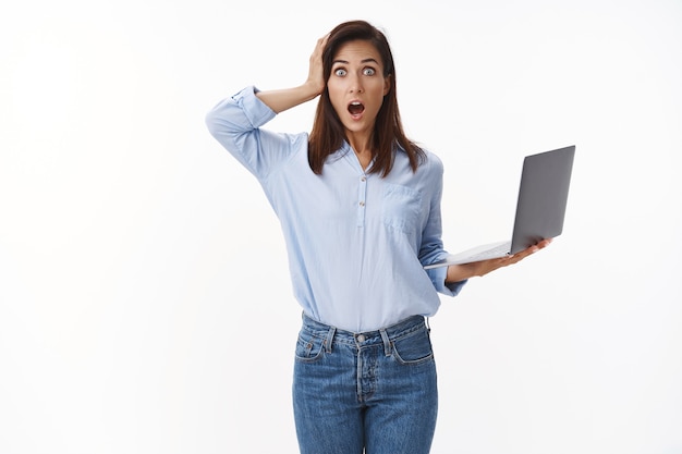 Troubled insecure female office newbie hold laptop, look concerned puzzled, panicking, open mouth gasping nervous grab head perplexed, distressed piles work, stand white wall anxious