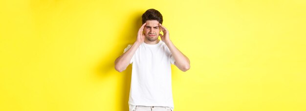 Troubled man touching head and grimacing from pain complaining on headache standing over yellow back