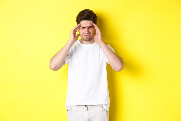 Free photo troubled man touching head and grimacing from pain, complaining on headache, standing over yellow background. copy space