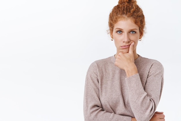 Troubled thoughtful gorgeous redhead female entrepreneur with messy hairbun touching lip and looking focused camera thinking during brainstorm trying create new ideas for project