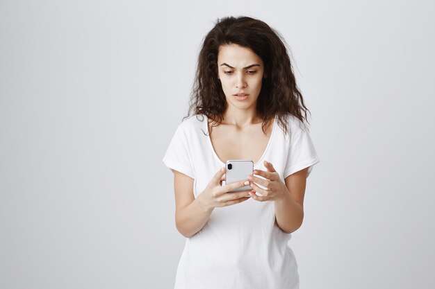 Обеспокоенная и обеспокоенная девушка смотрит на мобильный телефон с растерянным выражением лица