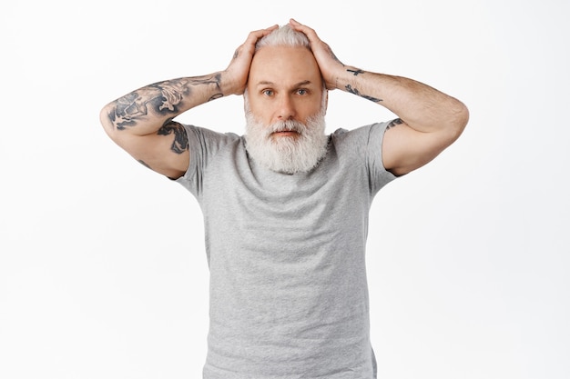 Free photo troubled and worried old man with tattoos, holding hands on head and look confused, panicking or feeling nervous about accident, standing alarmed and indecisive against white wall