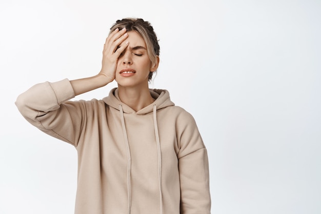 Troubled young woman, having headache, holding hand on head, facepalm gesture, standing distressed on white