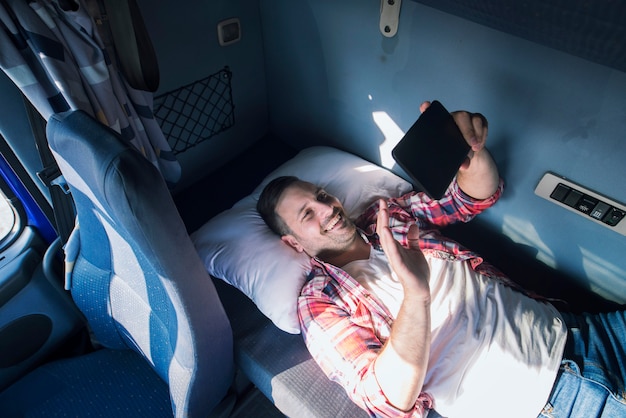 Free photo truck driver separated from his family lying on the bad of his truck cabin and waving to his wife and children via tablet computer