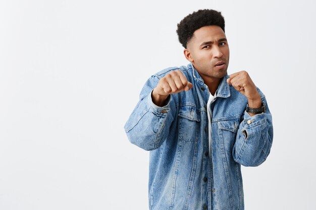 Try to beat me. Close up of young good-looking dark-skinned men with curly hair in white t shirt and denim jacket holding hands in fight pose with mean and aggressive expression.