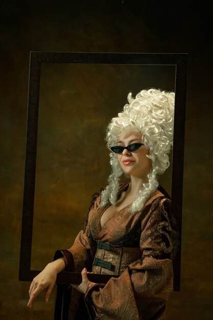 Trying on eyewear. Portrait of medieval young woman in vintage clothing with wooden frame on dark background. Female model as a duchess, royal person. Concept of comparison of eras, fashion, beauty.