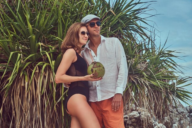 An ttractive couple, holds coconut with a tube, posing on a beach near palm bush, enjoys a vacation on a beautiful island.