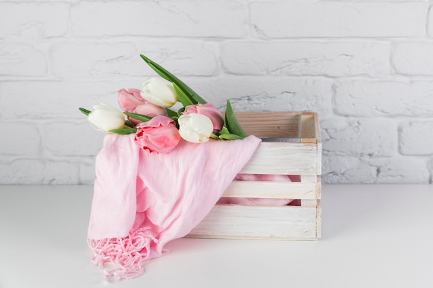 Tulips and pink scarf inside the wooden crate on desk against white brick wall