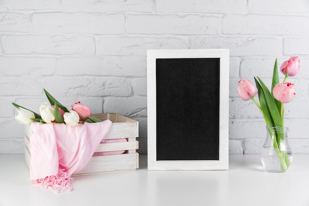 Tulips vase and crate near the blank black white border frame on desk against brick wall